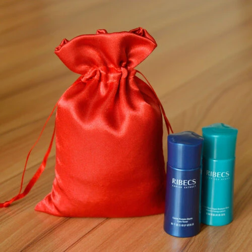 حقيبة حريرية مخصصة باللون الأحمر النابض بالحياة مع إغلاق برباط، مثالية لتخزين المجوهرات أو هدايا الزفاف، معروضة بجوار زجاجتين أسطوانيتين لمستحضرات التجميل تحملان علامة RIBECS.