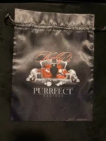 A black drawstring bag with a glossy finish featuring an orange and white graphic of a cat sitting on a throne surrounded by jewels, with the text “Felix” above and “PURRRFECT DESIRES” below.