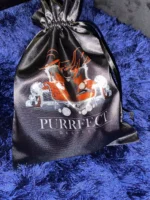 A black drawstring bag with a glossy finish, featuring an orange stylized text “Purrfect” above an image of two red high-heeled shoes with diamond-like embellishments. The shoes are placed on a reflective surface, and the background includes a blue furry texture.